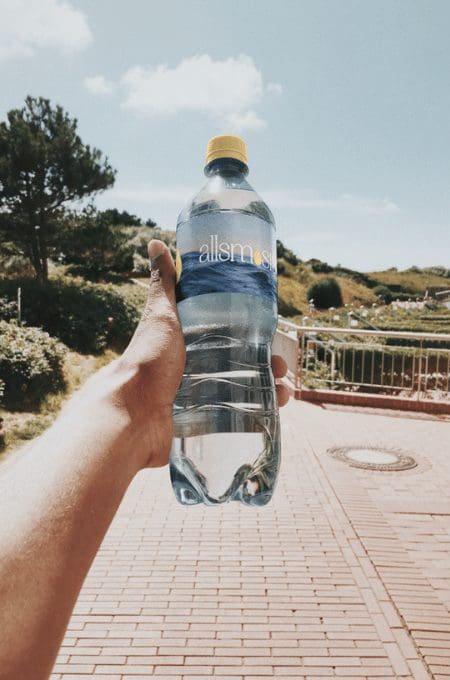 Hand holding a plastic water bottle labeled "allsmosis" outdoors on a sunny day, with a brick pathway and greenery in the background, conveying freshness.