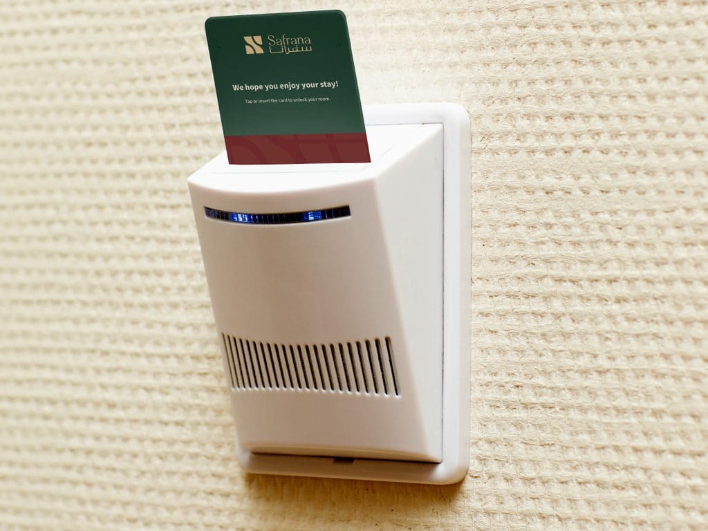 Hotel key card of Safrana in a wall slot with lights indicating power activation, set against a textured beige wall. The card conveys a welcoming message.
